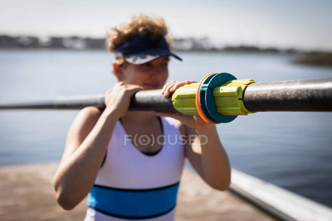 Vista frontal de cerca de una mujer remo caucásica llevando un remo en su hombro, caminando en un embarcadero en el sol sonriendo, enfoque selectivo - foto de stock