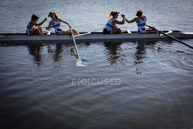 На березі видніється веслувальний колектив з чотирьох кавказьких жінок, які тренуються на річці, сидячи в мушлі на сонці, потискуючи руки і посміхаючись після веслування. — стокове фото