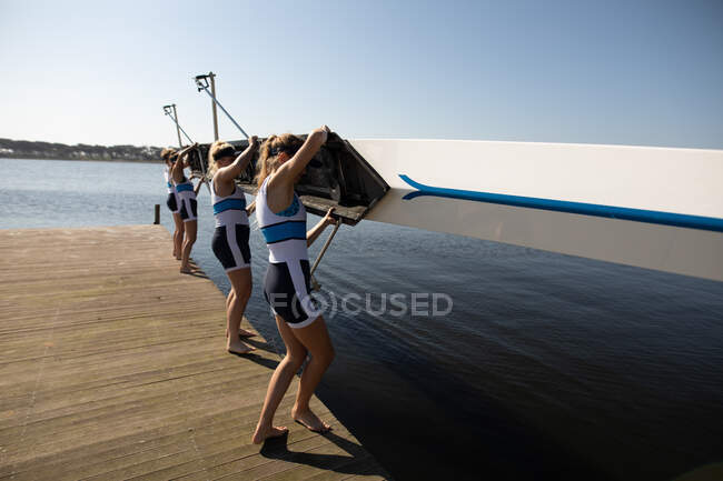 Vista lateral de uma equipe de remo de quatro mulheres caucasianas treinando no rio, em um molhe ao sol abaixando um barco na água antes de remar — Fotografia de Stock