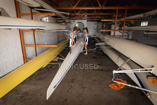 Vue latérale d'une équipe d'aviron de quatre femmes caucasiennes soulevant un bateau dans un hangar à bateaux, avant de le porter à la rivière pour s'entraîner — Photo de stock