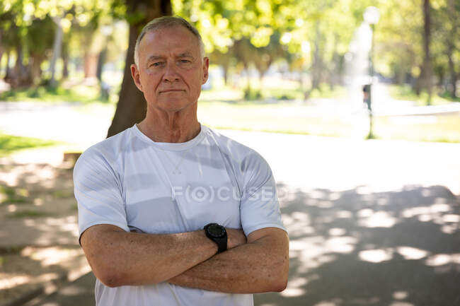 Porträt eines älteren kaukasischen Mannes, der es genießt, an einem sonnigen Tag in einem Park zu arbeiten und mit verschränkten Armen in die Kamera mit Bäumen im Hintergrund blickt — Stockfoto