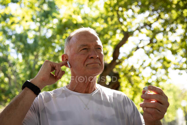 Niedrige Blickrichtung von einem älteren kaukasischen Mann, der an einem sonnigen Tag in einem Park trainiert, Kopfhörer aufsetzt und sich auf das Training vorbereitet — Stockfoto