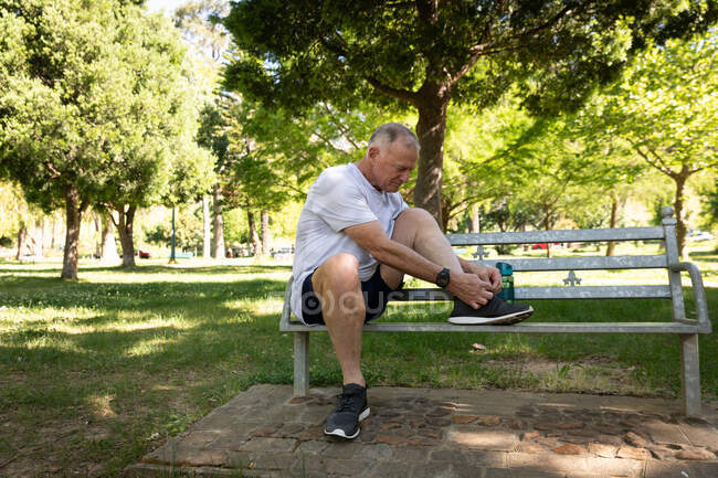 Vista lateral de un hombre mayor caucásico maduro haciendo ejercicio en un parque en un día soleado, sentado en un banco, atándose los cordones de los zapatos - foto de stock