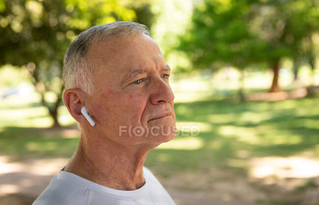 Vista lateral de un hombre mayor caucásico maduro haciendo ejercicio en un parque en un día soleado, con auriculares puestos, preparándose para hacer ejercicio - foto de stock