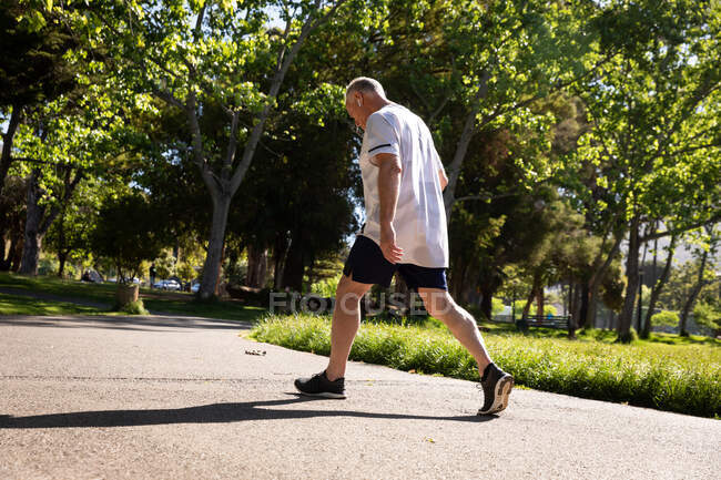 Low angle rear view of a athletic senior Caucasian man working out in a park, walking with earphones on — Stock Photo