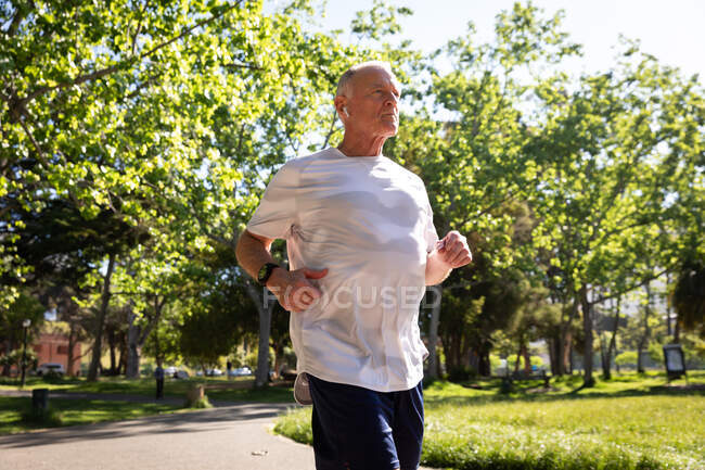 Low angle side view of a athletic senior Caucasian man working out in a park on a sunny day, running with earphones on — Stock Photo