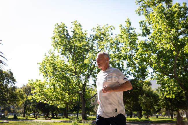 Side view of a athletic senior Caucasian man working out in a park on a sunny day, running with earphones on — Stock Photo