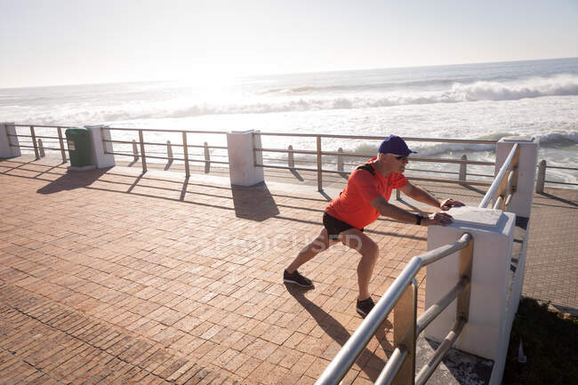 Side view of a mature senior Caucasian man working out on a promenade on a sunny day, stretching and leaning on balustrade — Stock Photo
