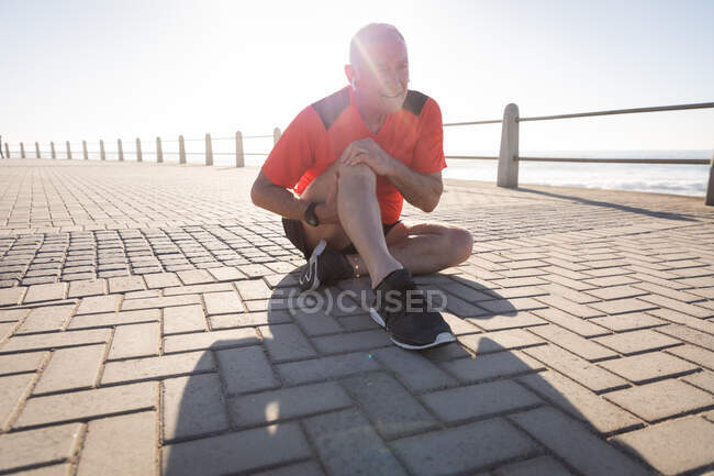 Vorderansicht eines älteren kaukasischen Mannes, der an einem sonnigen Tag mit blauem Himmel auf einer Promenade trainiert, sein Knie hält und mit Kopfhörern auf dem Bürgersteig sitzt — Stockfoto