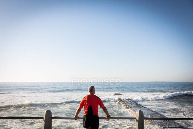Vista trasera de un hombre mayor caucásico maduro trabajando en un paseo marítimo en un día soleado con cielo azul, tomando un descanso, aferrándose a una balaustrada y admirando la vista - foto de stock