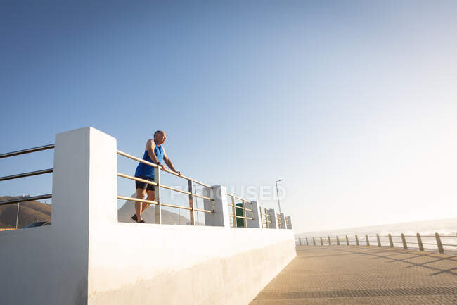 Niedrige Seitenansicht eines älteren kaukasischen Mannes, der an einem sonnigen Tag mit blauem Himmel auf einer Promenade trainiert, eine Pause einlegt, die Aussicht bewundert und sich an einer Brüstung festhält. — Stockfoto