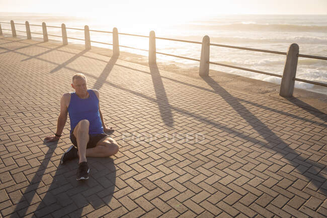 Вид сбоку на взрослого старшего кавказца, тренирующегося на набережной в солнечный день, отдыхающего, сидящего на тротуаре — стоковое фото