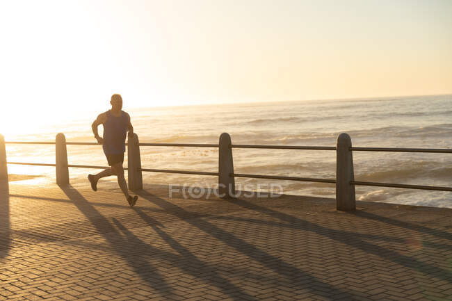 Вид сбоку на взрослого кавказца, тренирующегося на набережной в солнечный день, бегущего по морю — стоковое фото