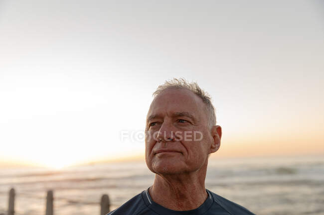 Vista frontal de un hombre mayor caucásico maduro haciendo ejercicio en un paseo marítimo, admirando la vista durante la puesta del sol - foto de stock