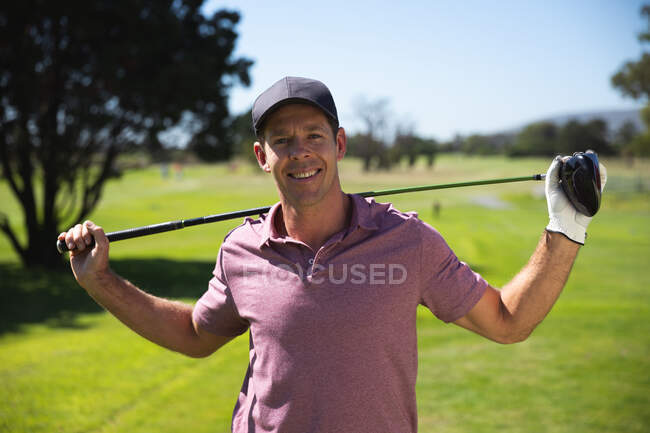 Портрет кавказького чоловіка на полі для гольфу в сонячний день з блакитним небом, тримаючи гольф клуб через плечі, посміхаючись до камери — стокове фото