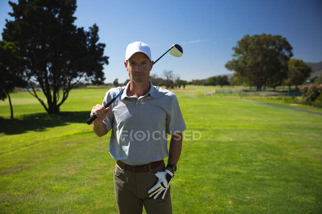 Retrato de um homem caucasiano em um campo de golfe em um dia ensolarado com céu azul, segurando um taco de golfe em seu ombro, sorrindo para a câmera — Fotografia de Stock