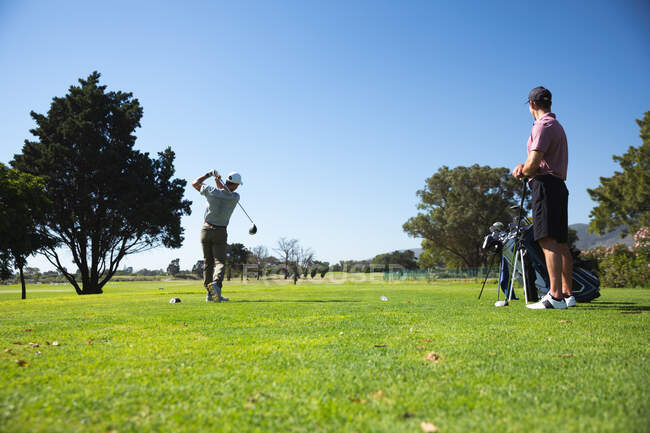 Vue arrière d'un deux hommes caucasiens sur un terrain de golf par une journée ensoleillée avec ciel bleu, l'un frappant une balle et l'autre debout et regardant — Photo de stock