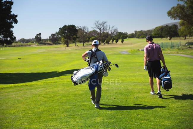 Vista trasera de dos hombres caucásicos en un campo de golf en un día soleado con cielo azul, caminando, llevando bolsas de golf - foto de stock