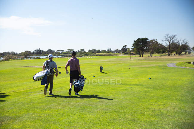 Вид сзади на двух кавказских мужчин на поле для гольфа в солнечный день с голубым небом, гуляющих, несущих сумки для гольфа — стоковое фото