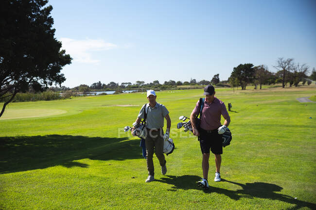 Перед очима двох кавказьких чоловіків на полі для гольфу в сонячний день з блакитним небом, йдучи, несучи мішки для гольфу. — стокове фото