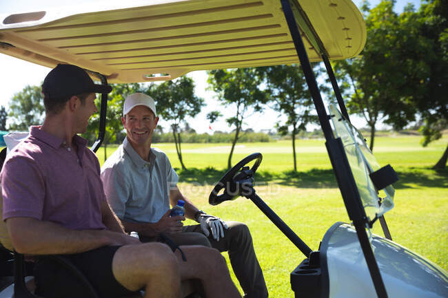 Vista lateral de dos hombres caucásicos en un campo de golf en un día soleado, sentado en un carrito de golf, hablando - foto de stock