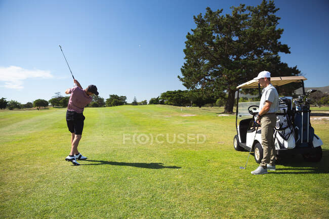 Vista posteriore di due uomini caucasici in un campo da golf in una giornata di sole con cielo blu, uno che colpisce una palla e l'altro in piedi e guardare — Foto stock