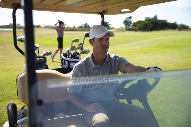 Вид спереди на кавказца на поле для гольфа в солнечный день с голубым небом, за рулем гольф-кара, другой человек играет в гольф на заднем плане — стоковое фото