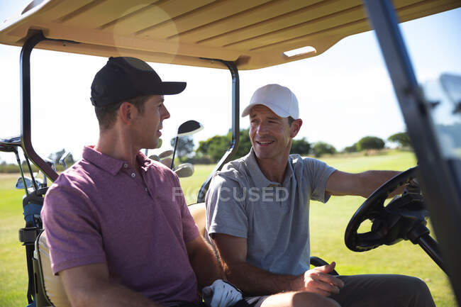 Вид спереди на белых мужчин на поле для гольфа в солнечный день с голубым небом, сидящих в гольф-каре, разговаривающих — стоковое фото