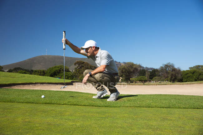 Вид сбоку на кавказца на поле для гольфа в солнечный день с голубым небом, стоящего на коленях, держащего клюшку для гольфа — стоковое фото