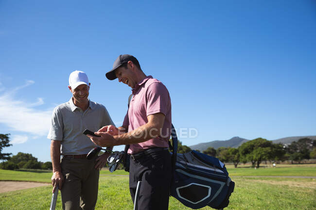 Вид спереди на двух белых мужчин на поле для гольфа в солнечный день с голубым небом, с помощью смартфона, улыбающихся — стоковое фото
