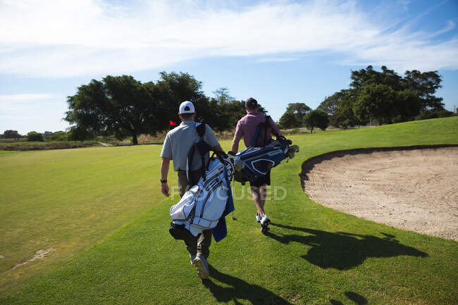 Rückansicht zweier kaukasischer Männer auf einem Golfplatz an einem sonnigen Tag mit blauem Himmel, beim Gehen, Golftaschen tragend — Stockfoto