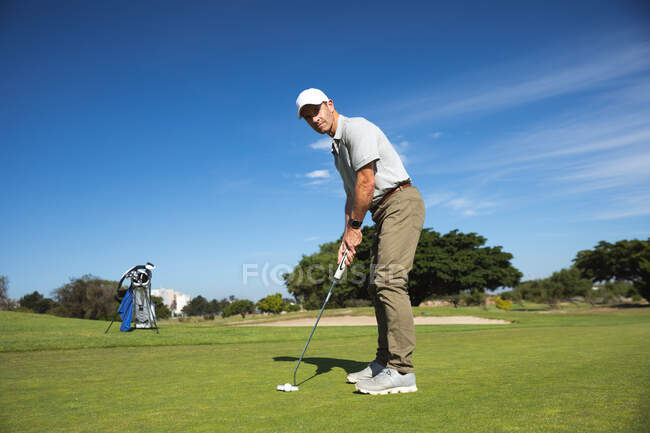 Vista lateral de un hombre caucásico en un campo de golf en un día soleado con cielo azul, golpeando una pelota de golf - foto de stock