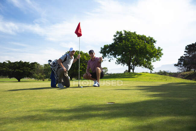 Vista frontal de dos hombres caucásicos en un campo de golf en un día soleado con cielo azul, en cuclillas, sosteniendo una bandera - foto de stock