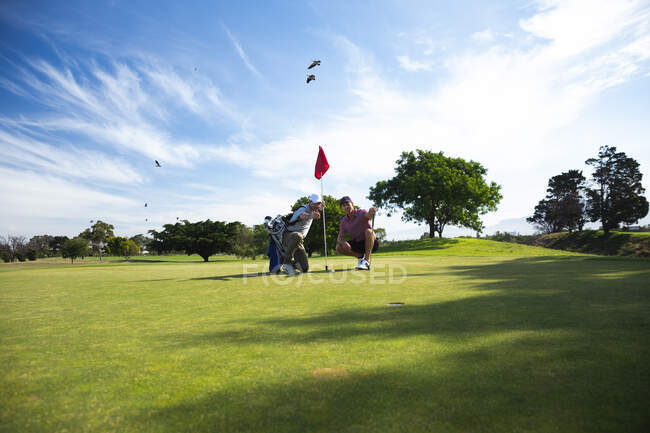 Vista frontale di due uomini caucasici in un campo da golf in una giornata di sole con cielo blu, accovacciato, con una bandiera in mano — Foto stock