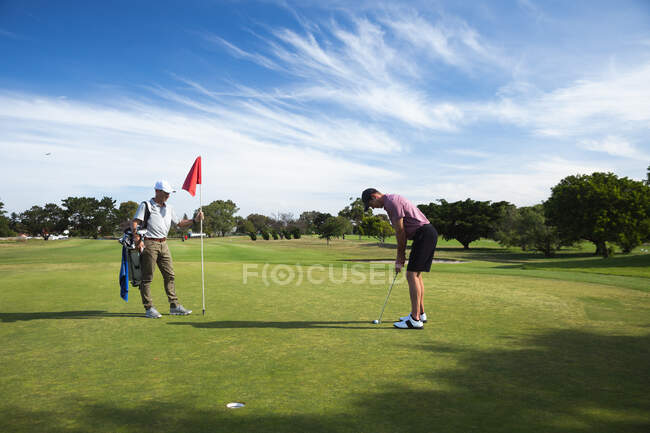 Vista lateral de dos hombres caucásicos en un campo de golf en un día soleado con cielo azul, golpeando una pelota de golf - foto de stock