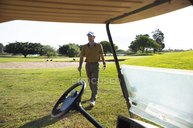 Frontansicht eines kaukasischen Mannes auf einem Golfplatz an einem sonnigen Tag, der einen Golfschläger in der Hand hält und auf einen Golfwagen zugeht — Stockfoto