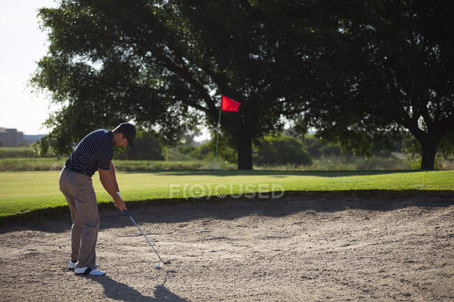 Задній вид на кавказького чоловіка на полі для гольфу в сонячний день, який б'є м'яч для гольфу в бункер. — стокове фото