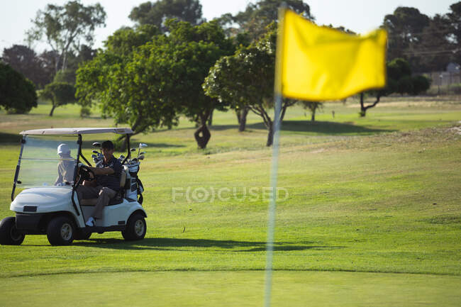 Вид спереди на двух белых мужчин на поле для гольфа в солнечный день, сидящих в гольф-каре с желтым флагом впереди — стоковое фото