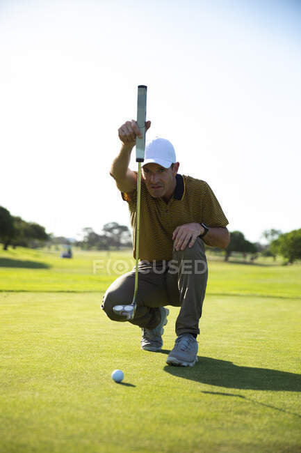 Vue de face d'un homme caucasien sur un terrain de golf, accroupi et vérifiant avant de frapper une balle dans le trou — Photo de stock