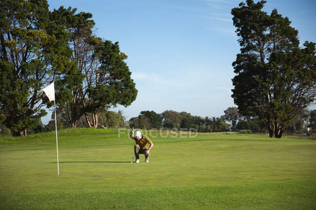 Вид спереди кавказца на поле для гольфа в солнечный день с голубым небом, сидящего на корточках и проверяющего перед ударом по мячу — стоковое фото