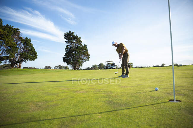 Vorderansicht eines kaukasischen Mannes auf einem Golfplatz an einem sonnigen Tag mit blauem Himmel, der einen Ball schlägt und zusieht, wie er ins Loch geht — Stockfoto