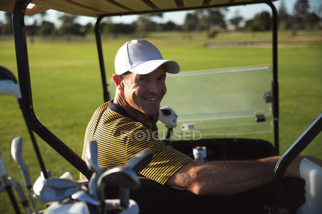 Portrait d'un homme caucasien sur un terrain de golf par une journée ensoleillée, assis dans une voiturette de golf, regardant la caméra sourire — Photo de stock