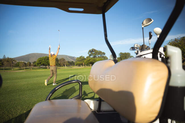 Parte trasera de un hombre caucásico en un campo de golf en un día soleado con cielo azul, de pie junto a un carrito de golf, sosteniendo sus manos hacia arriba en un gesto de triunfo - foto de stock