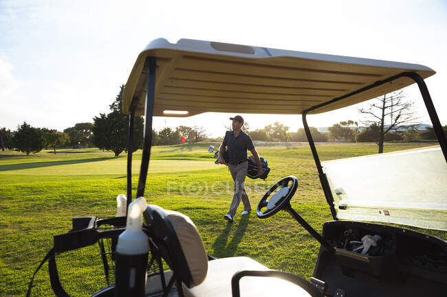 Vista lateral de un hombre caucásico en un campo de golf en un día soleado, caminando junto a un carrito de golf - foto de stock