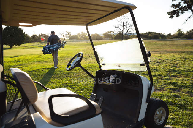 Погляд на кавказького чоловіка на поле для гольфу в сонячний день, коли він ходить з мішком для гольфу біля воза для гольфу. — стокове фото