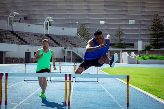 Front view of a Caucasian and a mixed race male athletes practicing at a sports stadium, racing each other on running track, hurdling — Stock Photo