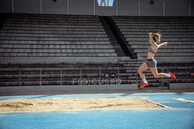Vue latérale d'une athlète blanche pratiquant dans un stade de sport, faisant un saut en longueur. — Photo de stock