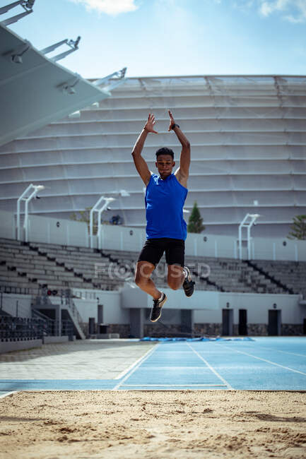 Vista frontal de un atleta masculino de raza mixta practicando en un estadio deportivo, haciendo un salto de longitud. - foto de stock