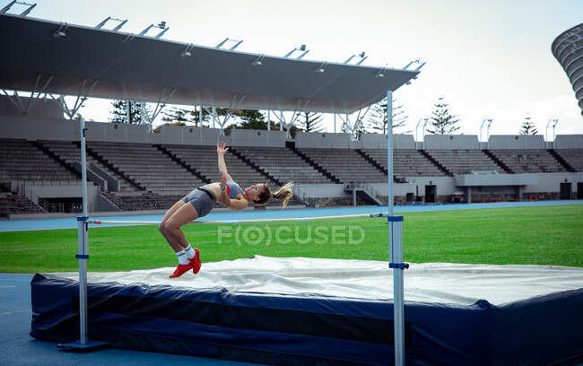 Vue latérale d'une athlète blanche pratiquant dans un stade de sport, faisant un saut en hauteur. — Photo de stock