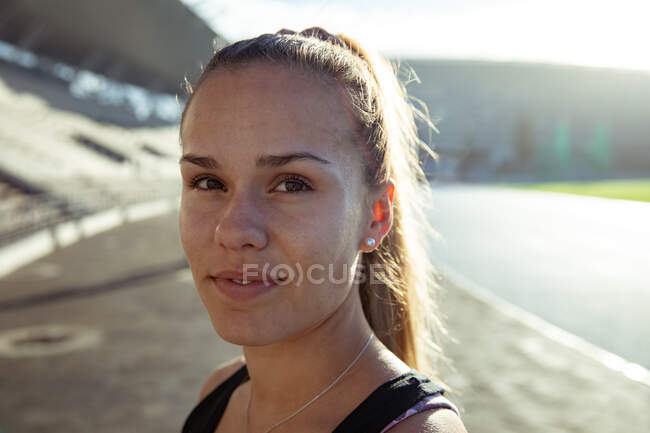 Retrato de una atleta blanca segura de sí misma con el pelo largo y rubio practicando en un estadio deportivo bajo el sol, mirando directamente a la cámara - foto de stock